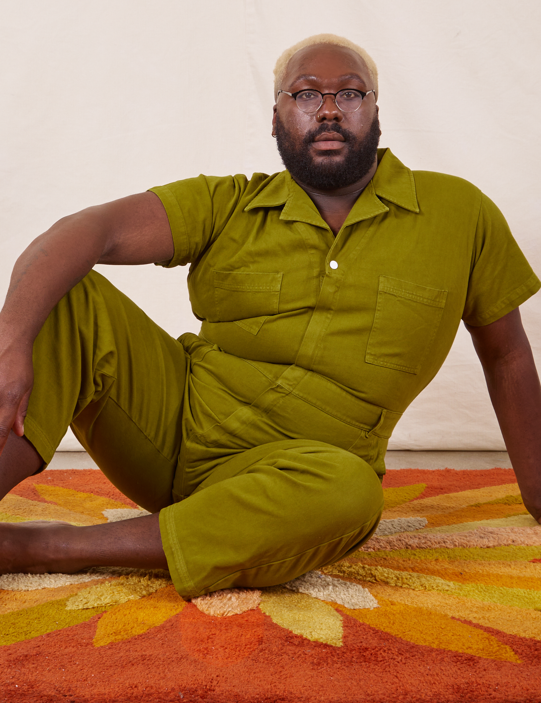 Elijah is sitting on a rug wearing Short Sleeve Jumpsuit in Olive Green