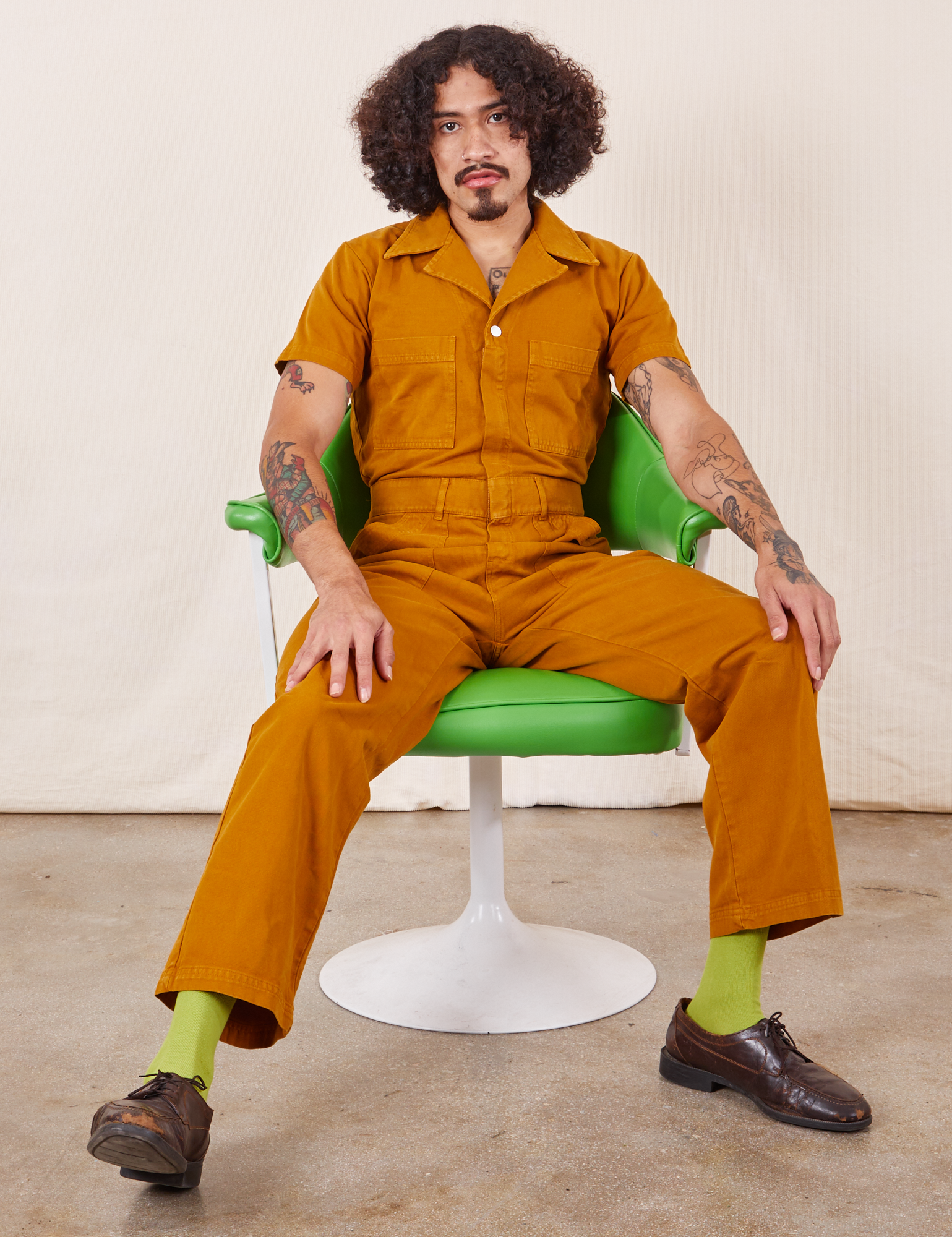 Jesse is sitting in a green vintage chair wearing Short Sleeve Jumpsuit in Spicy Mustard