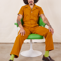 Jesse is sitting in a green vintage chair wearing Short Sleeve Jumpsuit in Spicy Mustard