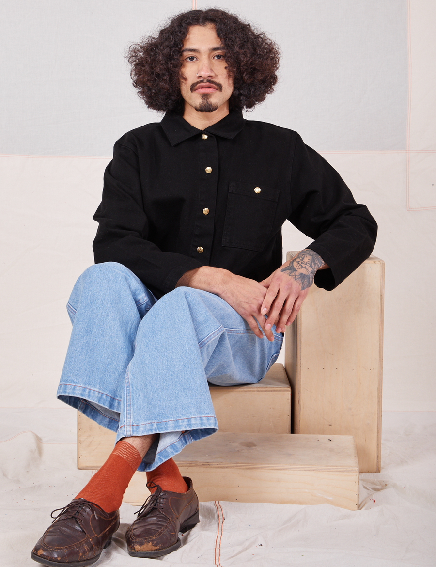 Jesse is sitting on a wooden crate. They are wearing a buttoned up Denim Work Jacket in Basic Black paired with light wash Sailor Jeans
