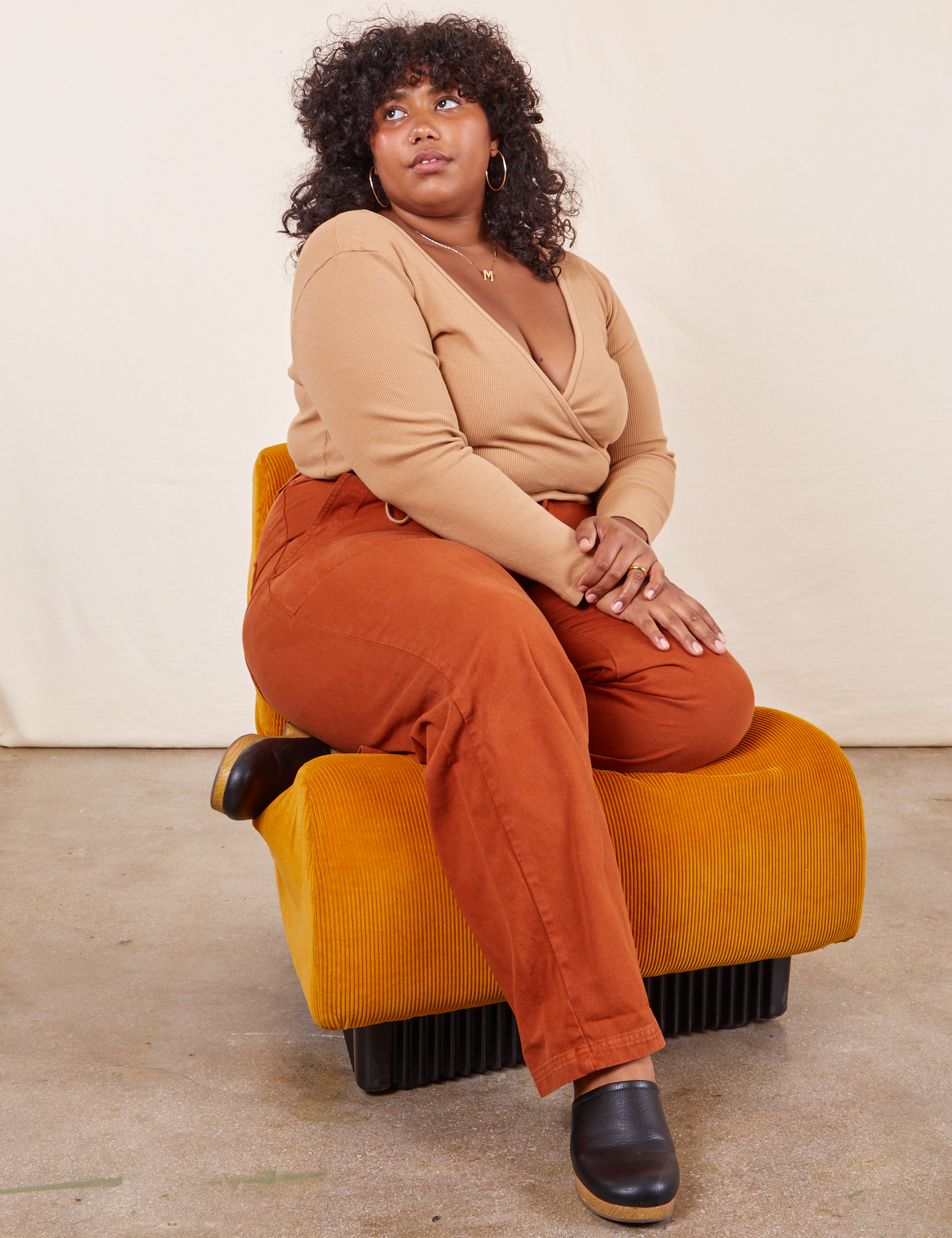 Morgan is sitting in an upholstered orange chair. She is wearing Work Pants in Burnt Terracotta paired with a tan Wrap Top