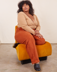 Morgan is sitting in an upholstered orange chair. She is wearing Work Pants in Burnt Terracotta paired with a tan Wrap Top