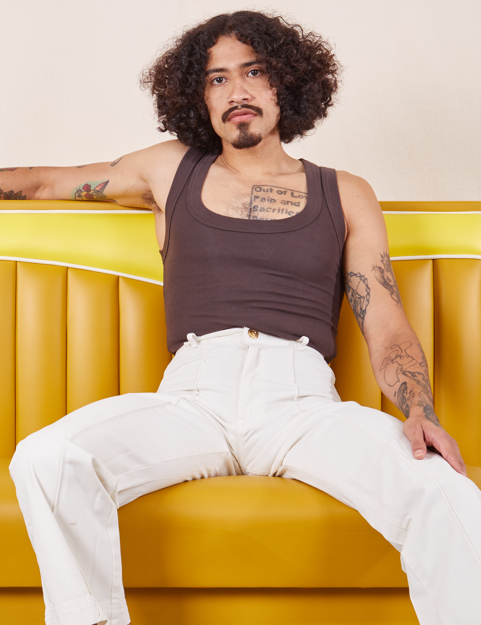 Jesse is wearing Western Pants in Vintage Tee Off-White and espresso brown Tank Top. They are sitting in a vinyl yellow dining booth.