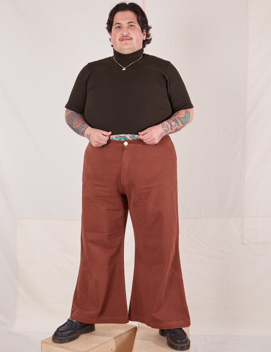 Sam is standing on two wooden crates wearing 1/2 Sleeve Essential Turtleneck in Espresso Brown and fudgesicle brown Bell Bottoms