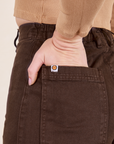 Back pocket close up of Western Pants in Espresso Brown. Worn by Alex with her hand in the pocket. Sun baby logo tag on top edge of pocket.
