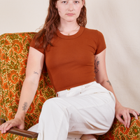 Alex is wearing Baby Tee in Burnt Terracotta sitting in a floral chair