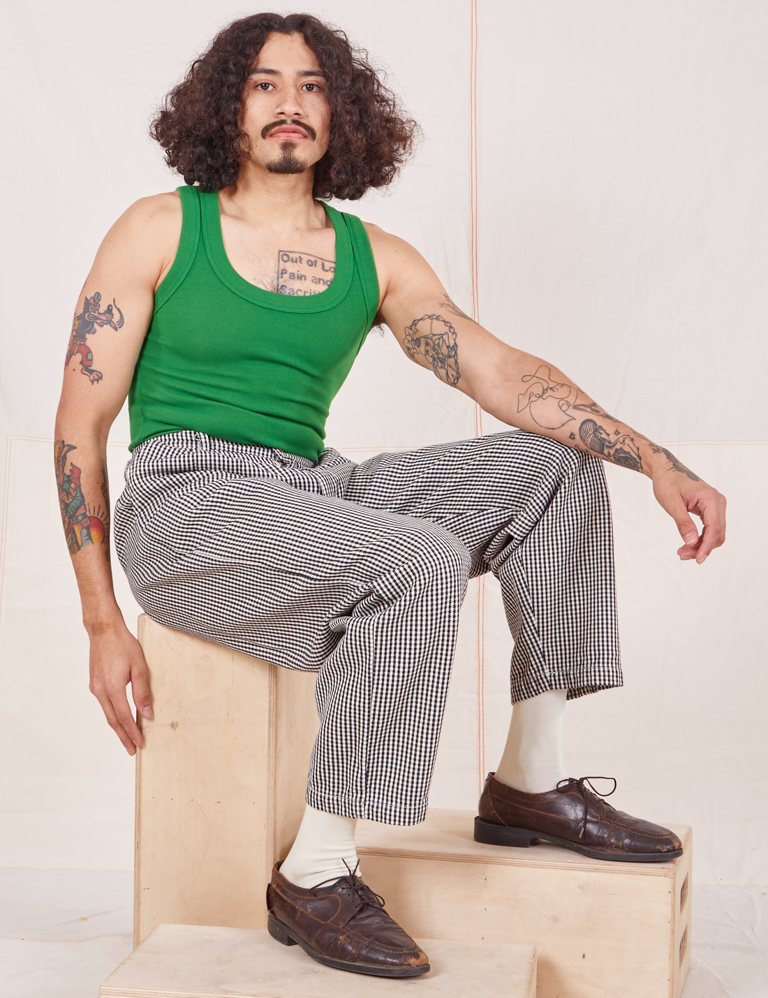 Jesse is sitting on a wooden crate wearing Checker Trousers in Black & White and forest green Tank Top