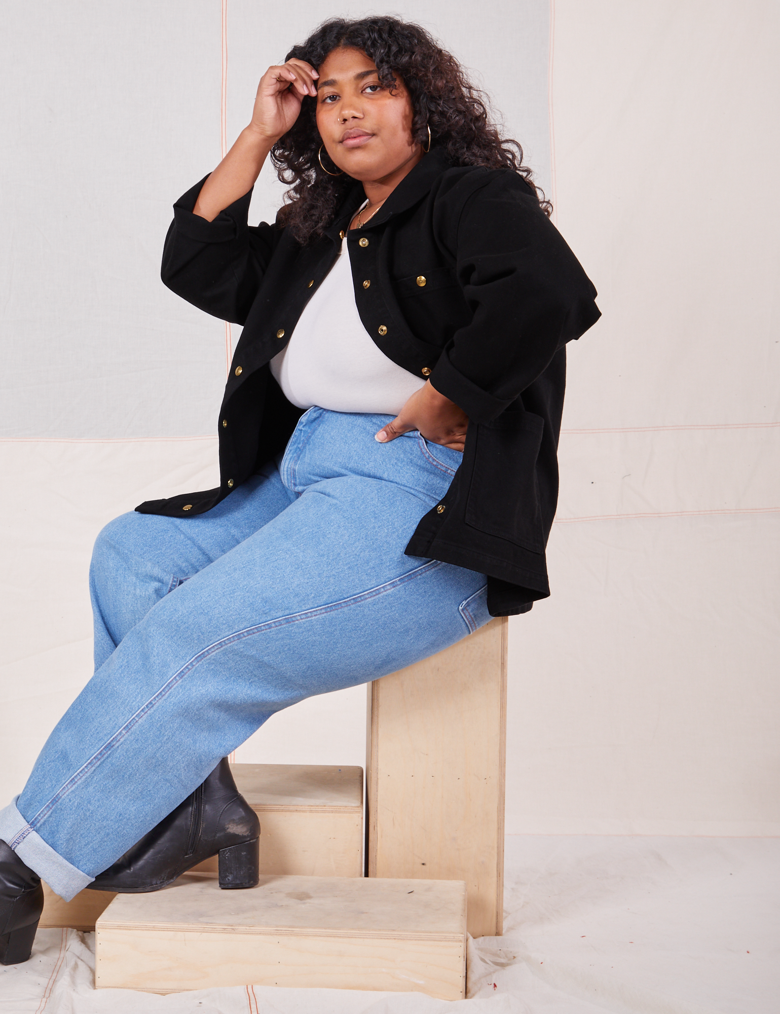 Morgan is sitting sideways on a wooden crate. She is wearing Denim Work Jacket in Basic Black, vintage off-white Baby Tee and light wash Frontier Jeans