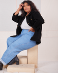 Morgan is sitting sideways on a wooden crate. She is wearing Denim Work Jacket in Basic Black, vintage off-white Baby Tee and light wash Frontier Jeans