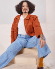 Jesse is sitting on a wooden crate. They are wearing Denim Work Jacket in Burnt Terracotta and light wash Sailor Jeans