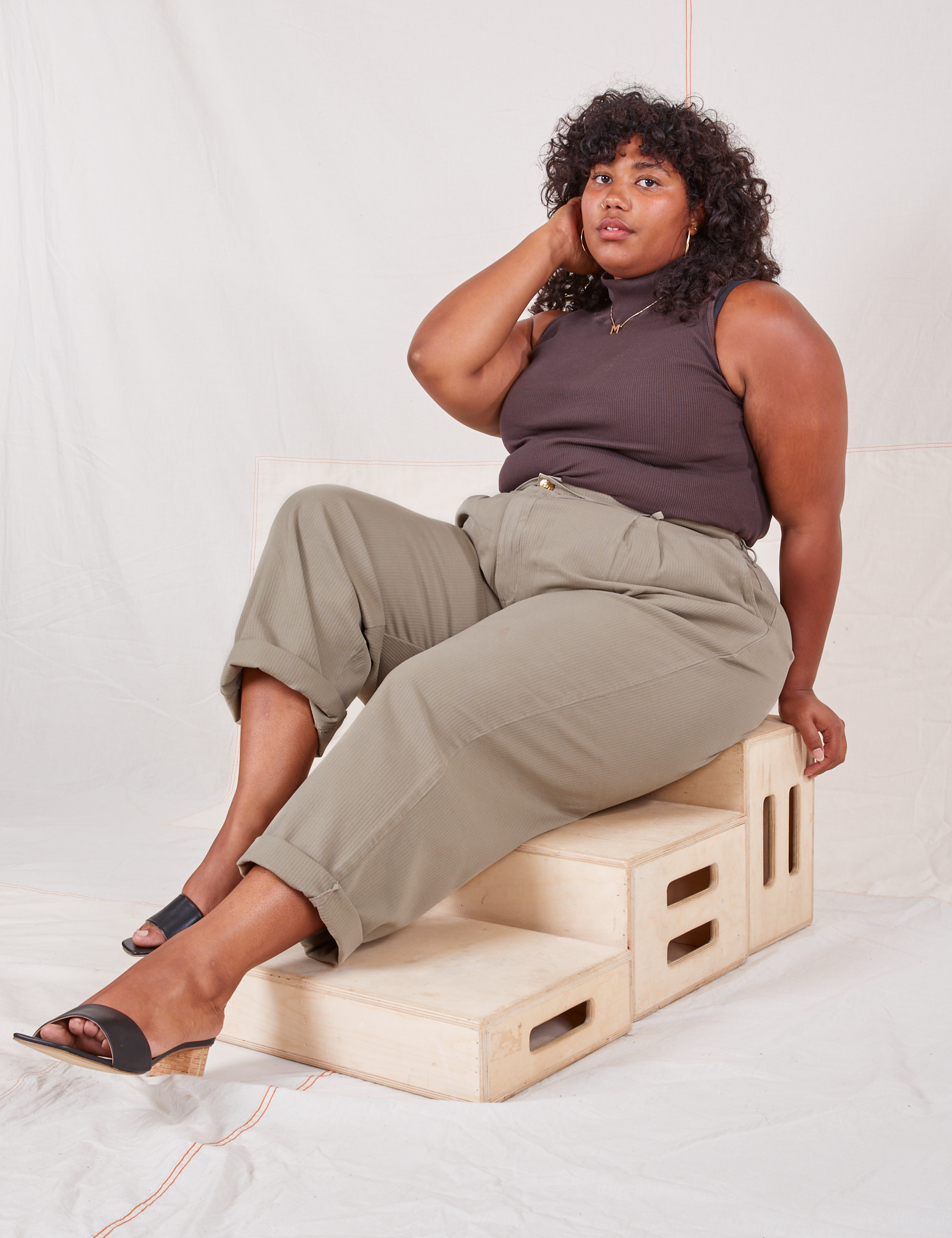 Heritage Trousers in Khaki Grey on Morgan sitting on wooden crates wearing espresso brown Sleeveless Essential Turtleneck