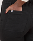 Back pocket close up of Organic Work Pants in Basic Black. Reece has her hand in the pocket.