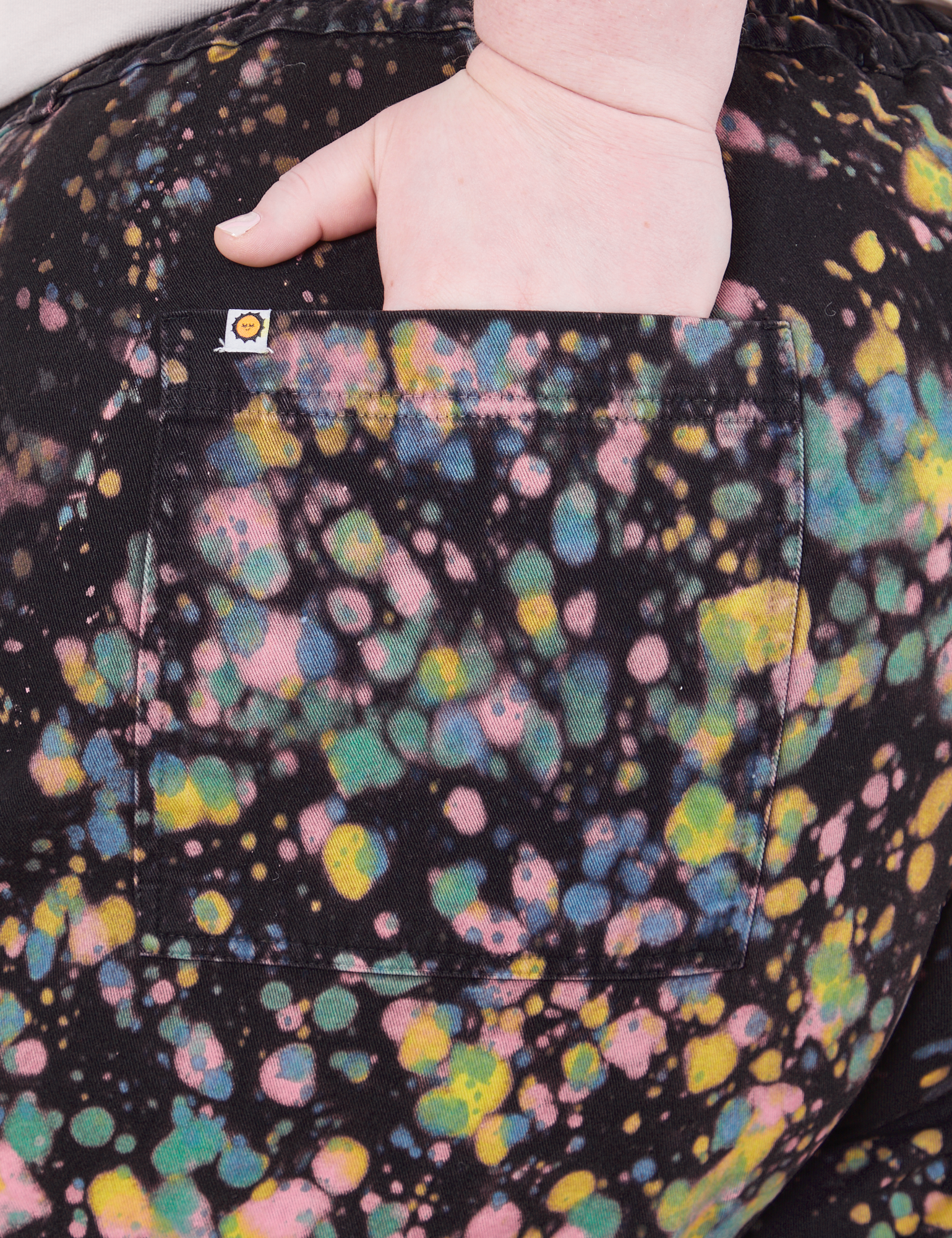Marble Paint Splatter Work Pants back pocket close up on Catie with hand in pocket. Sun baby logo tag on pocket