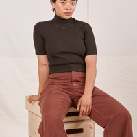 Mika is sitting on a stack of wooden crates wearing 1/2 Sleeve Essential Turtleneck in Espresso Brown and fudgesicle brown Bell Bottoms