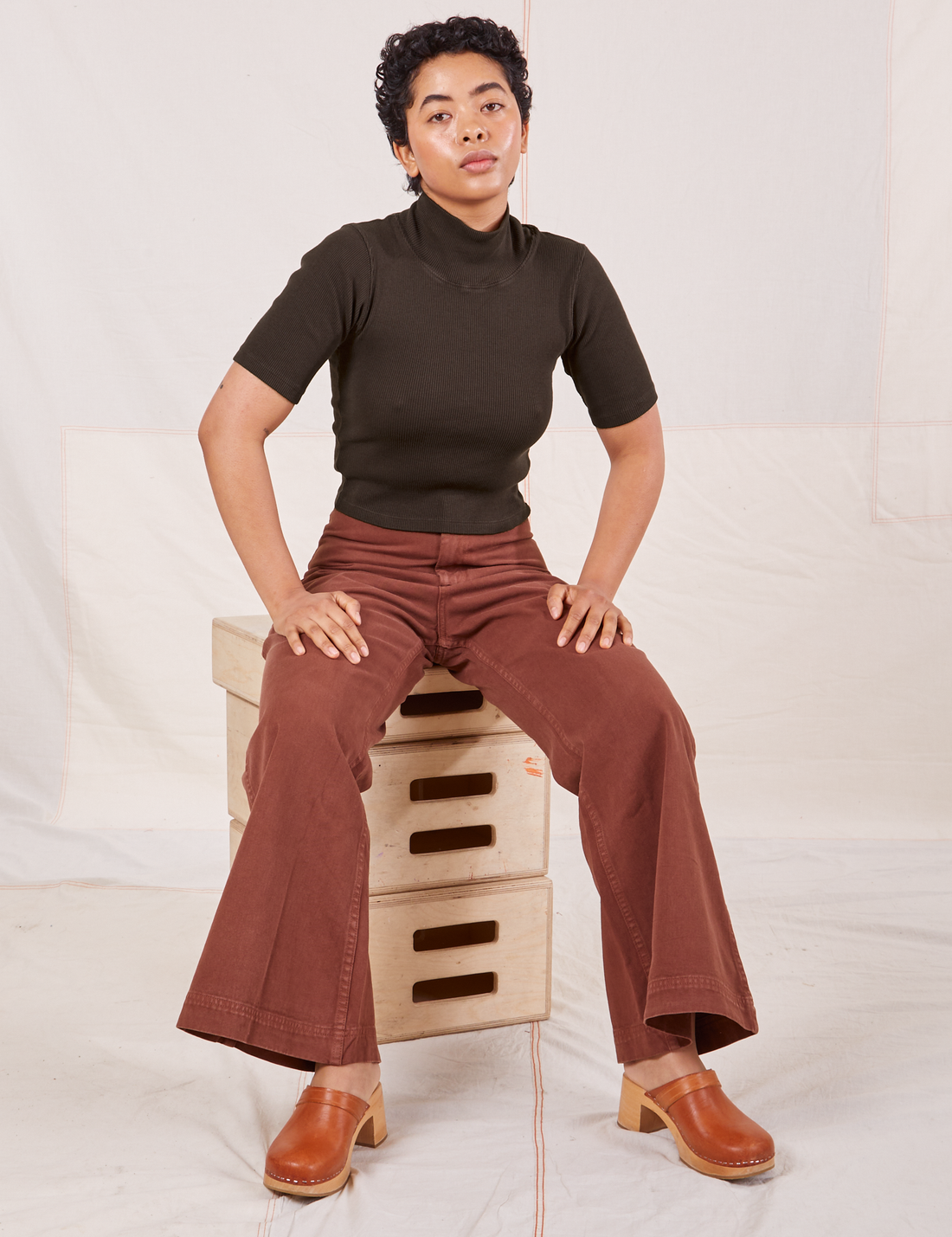 Mika is sitting on a stack of wooden crates wearing 1/2 Sleeve Essential Turtleneck in Espresso Brown and fudgesicle brown Bell Bottoms