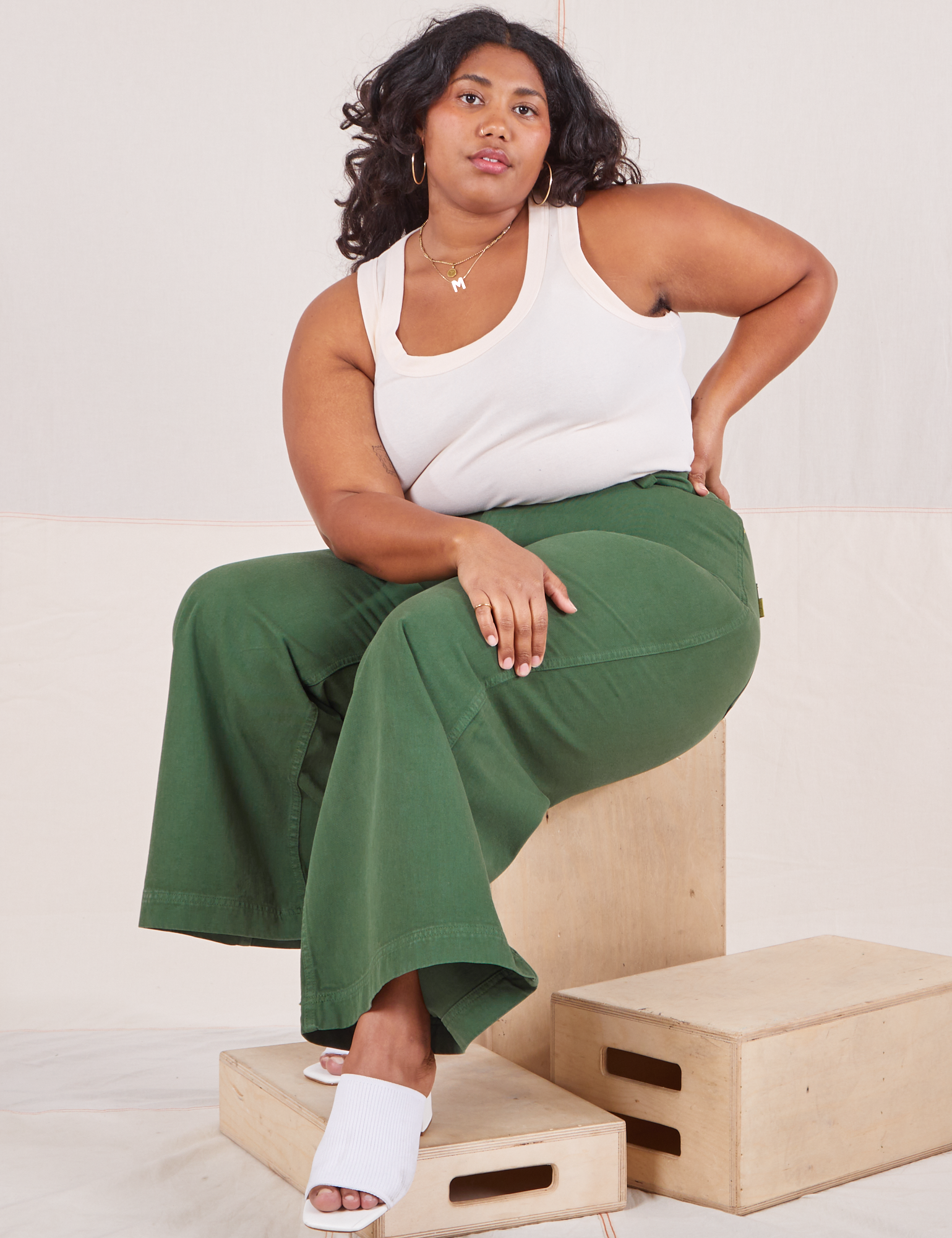 Morgan is sitting on a wooden crate wearing Bell Bottoms in Dark Emerald Green and vintage off-white Tank Top