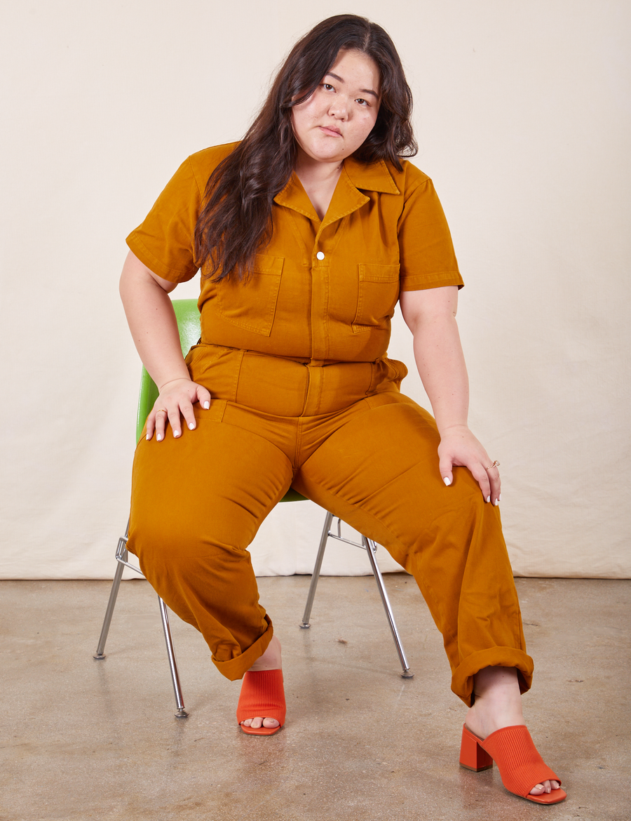 Ashley is sitting on a chair wearing Short Sleeve Jumpsuit in Spicy Mustard