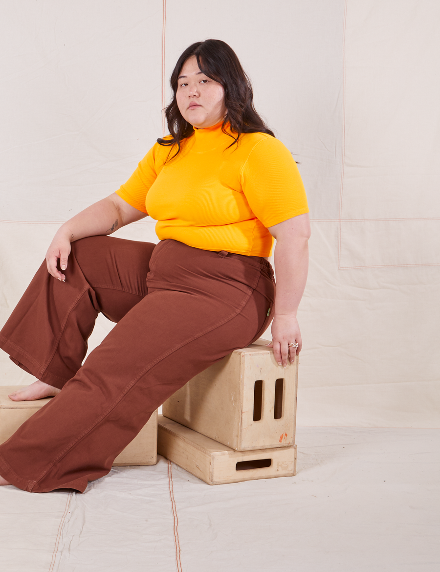 Ashley is sitting on a wooden crate wearing 1/2 Sleeve Essential Turtleneck in Sunshine Yellow and fudgesicle brown Bell Bottoms