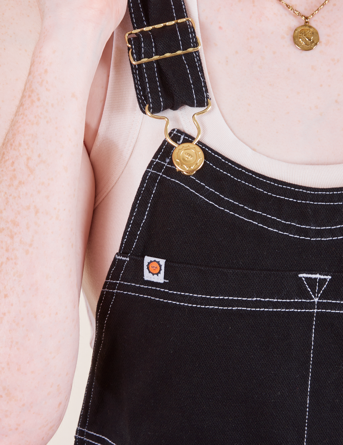 Original Overalls in Basic Black front strap close up featuring gold sun baby button and contrast white stitching