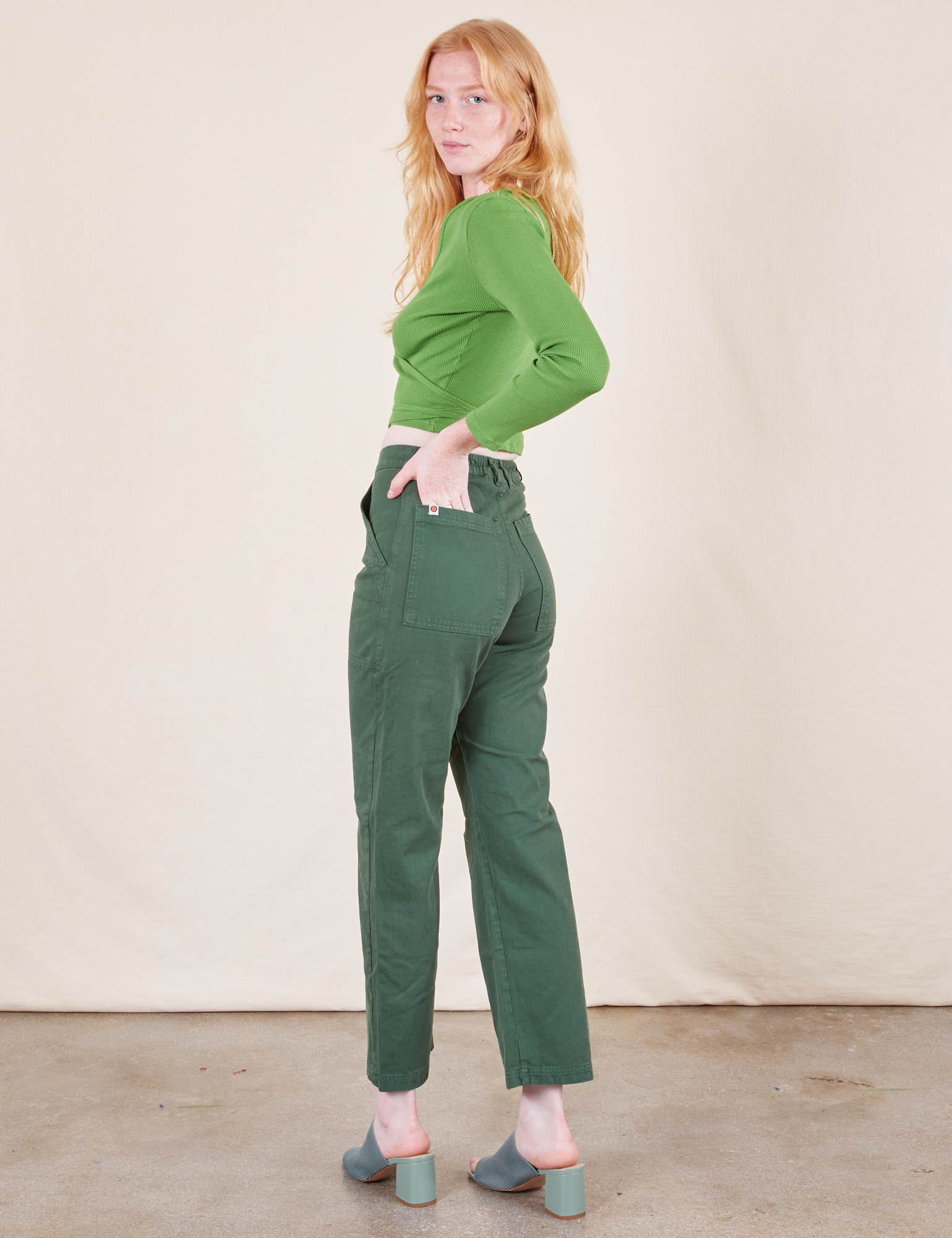 Angled back view of Work Pants in Dark Emerald Green and bright olive Wrap Top worn by Margaret